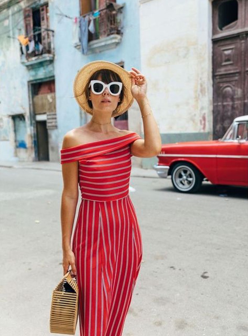 A woman in a red striped dress wearing sunglasses and a hat,  holding a bag, walking in the street.
